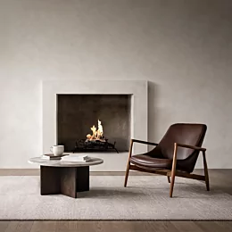 Modern minimalist living room featuring a built-in fireplace with a clean-burning flame set in a recessed concrete surround. In front, a round stone coffee table with books and a white mug sits on a textured rug beside a mid-century style brown leather lounge chair with wooden arms, creating a warm, contemporary interior setting.